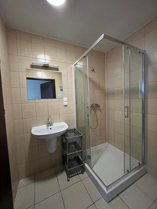 Łazienka w segmencie jednosobowym w Domu Studenta Fama. Jasne pomieszczenie z beżowymi kafelkami i beżową podłogą. Po prawej stronie znajduje się kabina prysznicowa, a po lewej zlew z lustrem ściennym i lampą nad nim. Obok kabiny prysznicowej stoi szafka na akcesoria łazienkowe i kosmetyki.