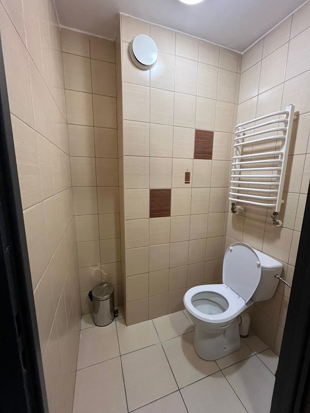 Toaleta w segmencie jednosobowym w Domu Studenta Fama. Jasne pomieszczenie z beżowymi kafelkami i beżową podłogą. Po prawej stronie znajduje się muszla klozetowa,nad nią umieszczony jest grzejnik. Po lewej kosz na śmieci.