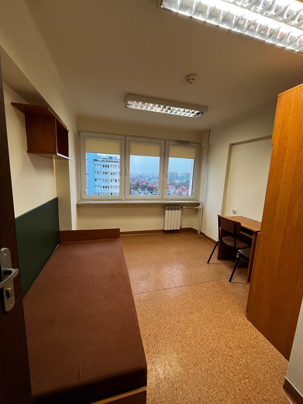 Jednoosobowy pok&oacute;j w domu studenta FAMA. Wnętrze jest proste i funkcjonalne: po prawej stronie stoi ł&oacute;żko z brązowym materacem, po lewej znajduje się biurko z krzesłem oraz wisząca p&oacute;łka. Na końcu pokoju są duże okna z widokiem na pobliskie bloki. Pomieszczenie oświetlają dwie sufitowe lampy jarzeniowe.