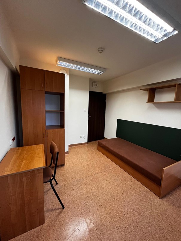 Jednoosobowy pok&oacute;j w domu studenta FAMA. Wnętrze jest proste i funkcjonalne: po prawej stronie stoi ł&oacute;żko z brązowym materacem, oraz wisząca p&oacute;łka ścienna, po lewej znajduje się biurko z jasnobrązowym krzesłem. Pomieszczenie oświetlają dwie sufitowe lampy jarzeniowe.