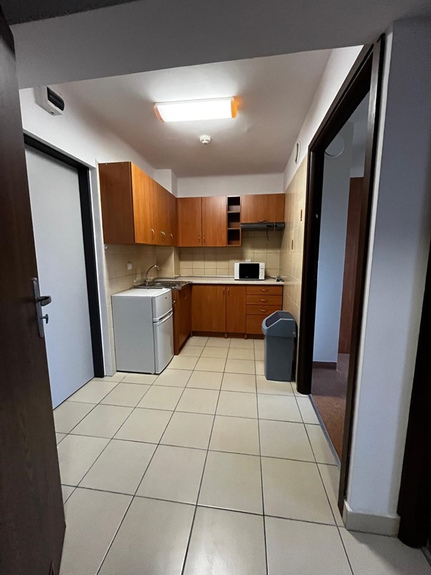 Kuchnia w domu studenta Fama z brązowymi szafkami i kremowym blatem; na blacie stoi mała biało-czarna mikrofal&oacute;wka. Wnętrze jest jasne i przestronne, z jasnobeżową podłogą i białymi ścianami; po lewej stronie znajduje się lod&oacute;wka, obok niej szafka ze zlewozmywakiem, a po prawej kosz na śmieci.