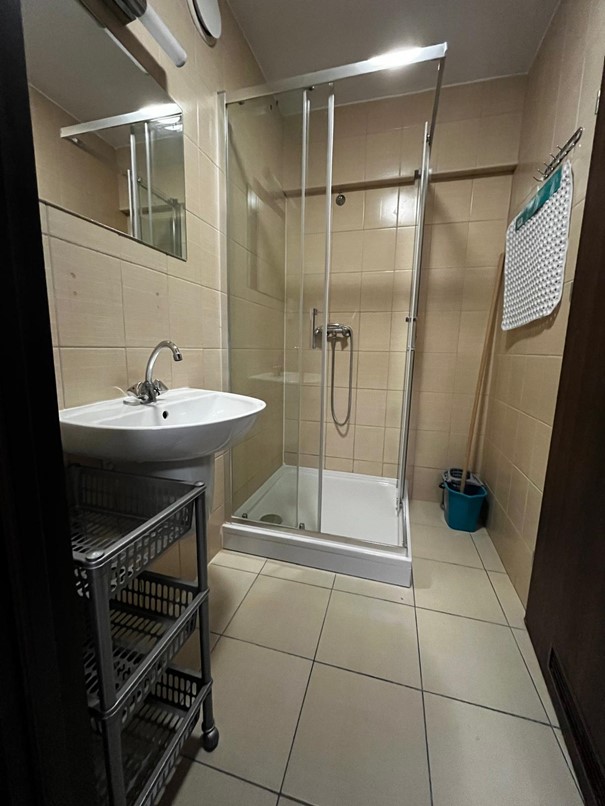 Łazienka w segmencie jednoosobowym w Domu Studenta Fama. Jasne pomieszczenie z beżowymi kafelkami i beżową podłogą. Po lewej stronie znajduje się kabina prysznicowa, a także zlew z lustrem ściennym i lampą nad nim. Obok zlewu stoi szafka na akcesoria łazienkowe i kosmetyki.