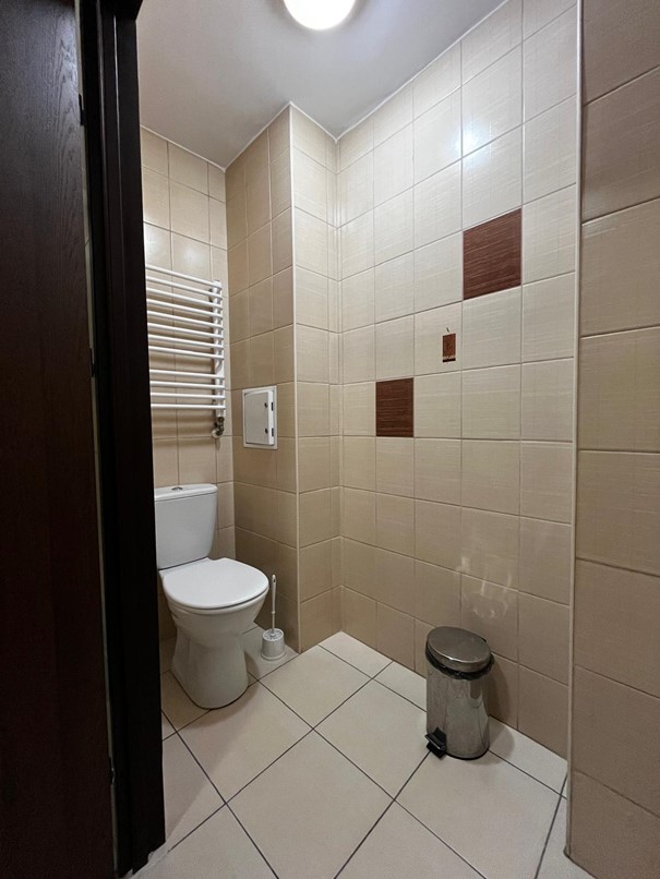 Toaleta w segmencie jednosobowym w Domu Studenta Fama. Jasne pomieszczenie z beżowymi kafelkami i beżową podłogą. Po lewej stronie znajduje się muszla klozetowa, nad kt&oacute;rą umieszczony jest grzejnik, a po prawej stoi kosz na śmieci.