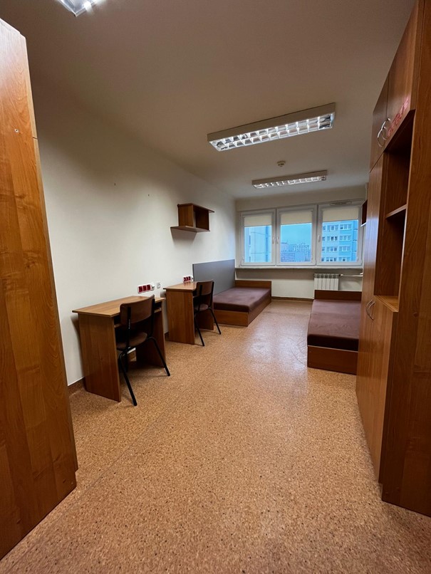 Pok&oacute;j dwuosobowy w domu studenta Fama z dużym oknem wyposażonym w białe rolety. W pomieszczeniu znajdują się dwie brązowe szafy, p&oacute;łki ścienne oraz dwa ł&oacute;żka w ciemnobrązowym kolorze. Podłoga jest jasnobrązowa, a wnętrze przestronne i dobrze oświetlone.