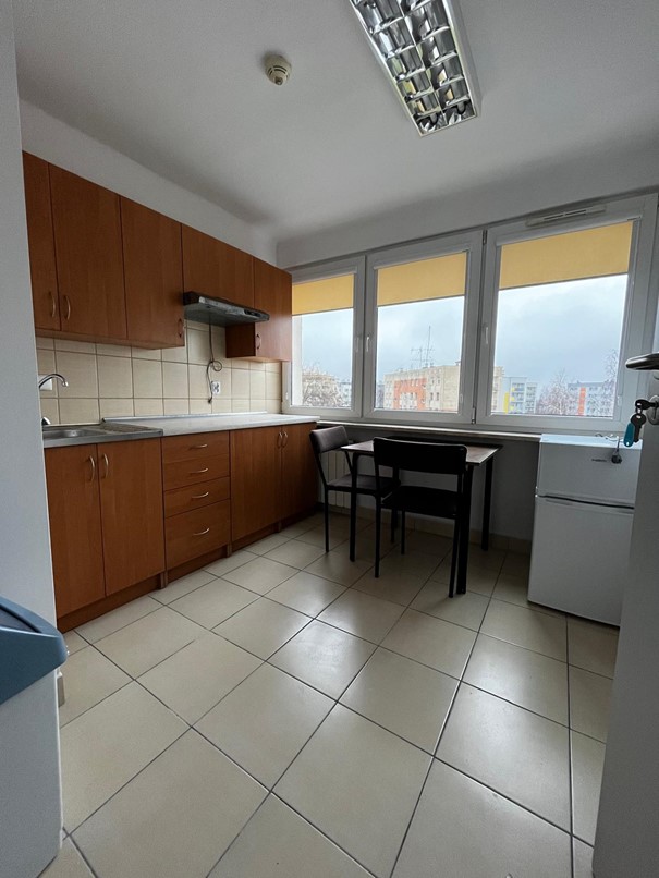 Przestronna kuchnia w segmencie dwuosobowym w domu studenta Fama, z jasnobrązowymi szafkami kuchennymi i jasnokremowymi kafelkami. Przy dużym oknie z roletami stoi st&oacute;ł oraz dwa ciemnobrązowe krzesła. Po prawej stronie znajduje się lod&oacute;wka, a po lewej kosz na śmieci.