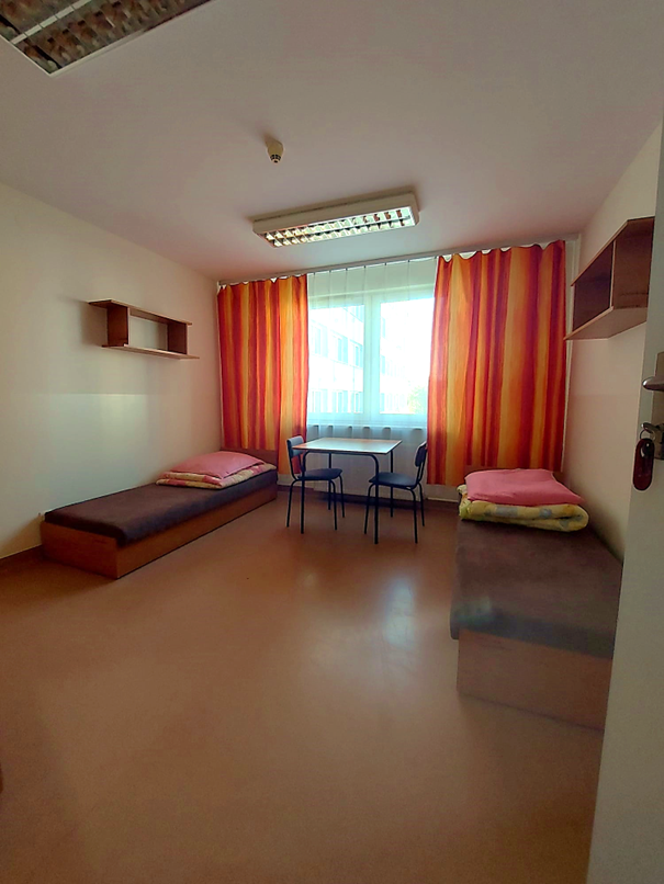 Zdjęcie przedstawia dwuosobowy pok&oacute;j w Domu Studenta Łącznik. W pomieszczeniu znajdują się dwa pojedyncze ł&oacute;żka z brązowymi materacami, st&oacute;ł z krzesłami w tym samym kolorze oraz p&oacute;łki ścienne wykonane z drewna w podobnym odcieniu. Na końcu pokoju znajduje się okno przez kt&oacute;re wpada naturalne światło.