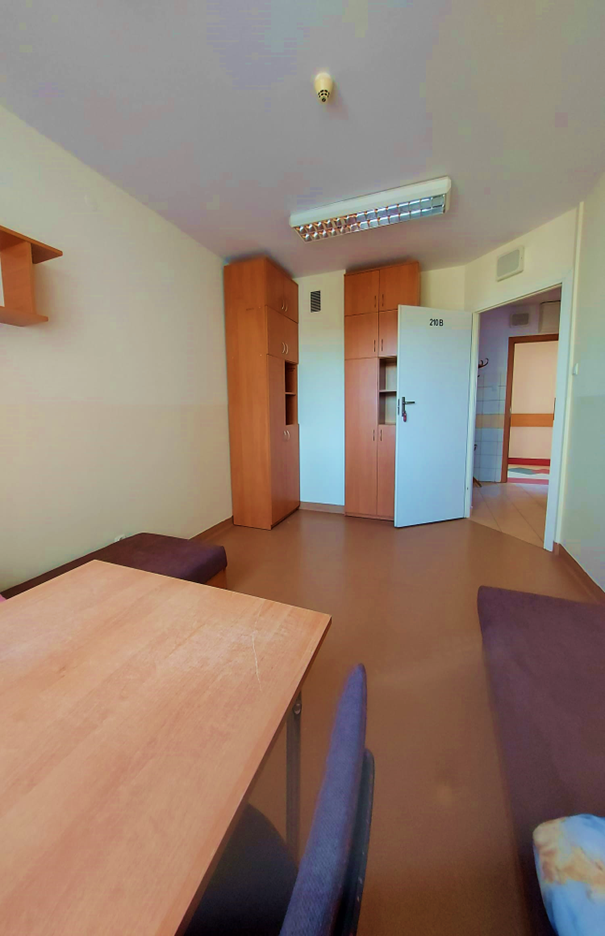 Zdjęcie przedstawia dwuosobowy pok&oacute;j w domu studenta Łącznik. W pomieszczeniu znajdują się dwa pojedyncze ł&oacute;żka z brązowymi materacami, st&oacute;ł z krzesłami w tym samym kolorze oraz duża szafa i p&oacute;łki ścienne wykonane z drewna w podobnym odcieniu. Na końcu pokoju znajduje się okno przez kt&oacute;re wpada naturalne światło.