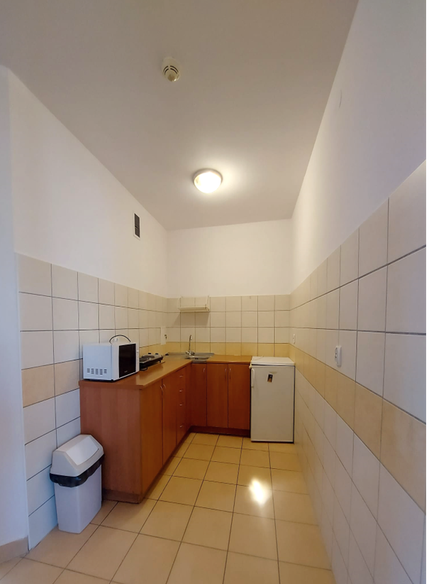 Zdjęcie przedstawia jasną kuchnię w akademiku Łącznik. Pomieszczenie ma jasne kafelki na podłodze i ścianach. Wzdłuż jednej ze ścian znajduje się blat kuchenny z szafkami i lod&oacute;wką. Kuchnia jest czysta i dobrze oświetlona.