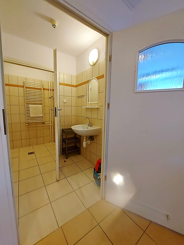 Zdjęcie przedstawia łazienkę w Domu Studenta Łącznik. Wnętrze wyłożone jest jasnymi beżowymi kafelkami. W pomieszczeniu znajduje się biała umywalka z lustrem oraz p&oacute;łka na akcesoria.