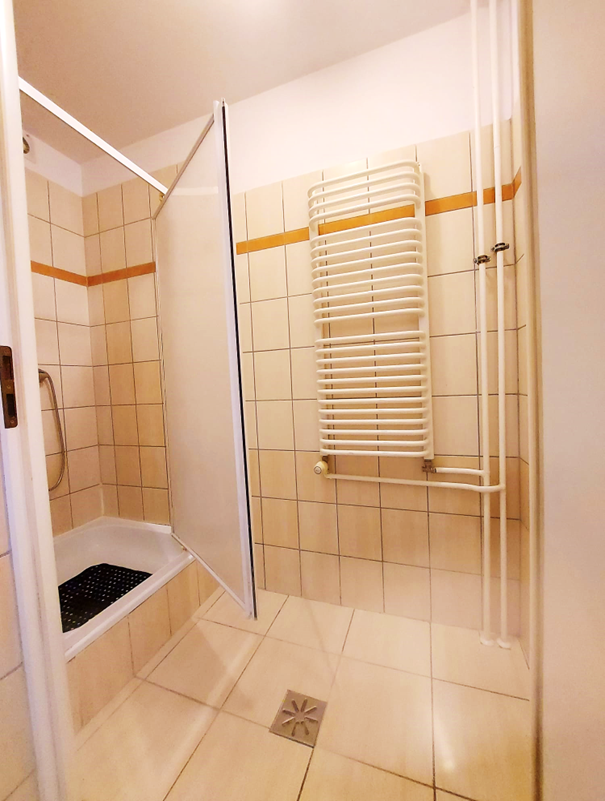 Zdjęcie przedstawia łazienkę w Domu Studenta Łącznik, Wnętrze wyłożone jest jasnymi płytkami. W pomieszczeniu znajduje się prysznic z uchylnymi matowymi drzwiami. Łazienka jest czysta, dobrze oświetlona i urządzona w prostym, funkcjonalnym stylu.