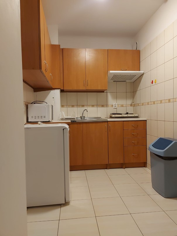 Zdjęcie przedstawia jasną kuchnię w Domu Studenta Melodia. W pomieszczeniu znajduje się zlewozmywak, płyta grzewcza, mikrofal&oacute;wka, kosz na śmieci, a także mała lod&oacute;wka oraz szafki kuchenne w kolorze drewna. Kuchnia jest czysta i dobrze oświetlona .