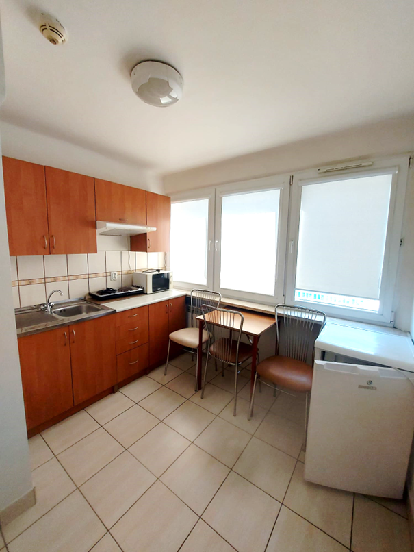 Zdjęcie przedstawia jasną kuchnię w Domu Studenta Melodia. W pomieszczeniu znajduje się zlewozmywak, płyta grzewcza, mikrofal&oacute;wka, mała lod&oacute;wka oraz szafki kuchenne w kolorze drewna. Przy oknie stoi st&oacute;ł z trzema krzesłami. Kuchnia jest czysta i dobrze oświetlona naturalnym światłem.