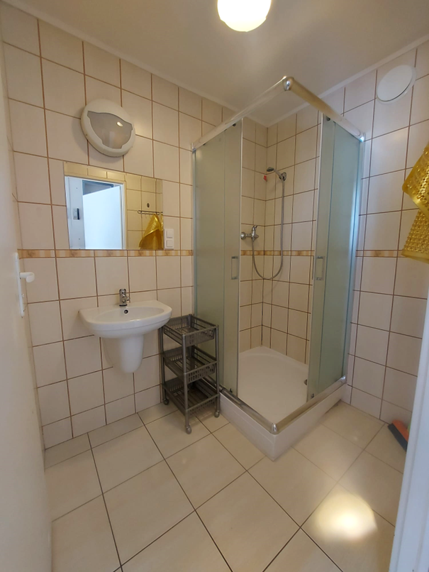 Zdjęcie przedstawia łazienkę w Domu Studenta Melodia, Wnętrze wyłożone jest jasnymi płytkami. W pomieszczeniu znajduje się prysznic z przesuwnymi, matowymi drzwiami, umywalka z lustrem oraz plastikowy stojak z p&oacute;łkami. Łazienka jest czysta, dobrze oświetlona i urządzona w prostym, funkcjonalnym stylu.