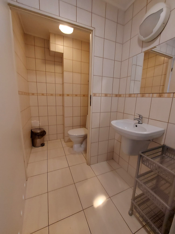 Jasna łazienka z toaletą w Domu Studenta Melodia. W pomieszczeniu znajduje się umywalka z lustrem i plastikowy regał na akcesoria. Łazienka jest wyłożona beżowo-brązowymi płytkami.