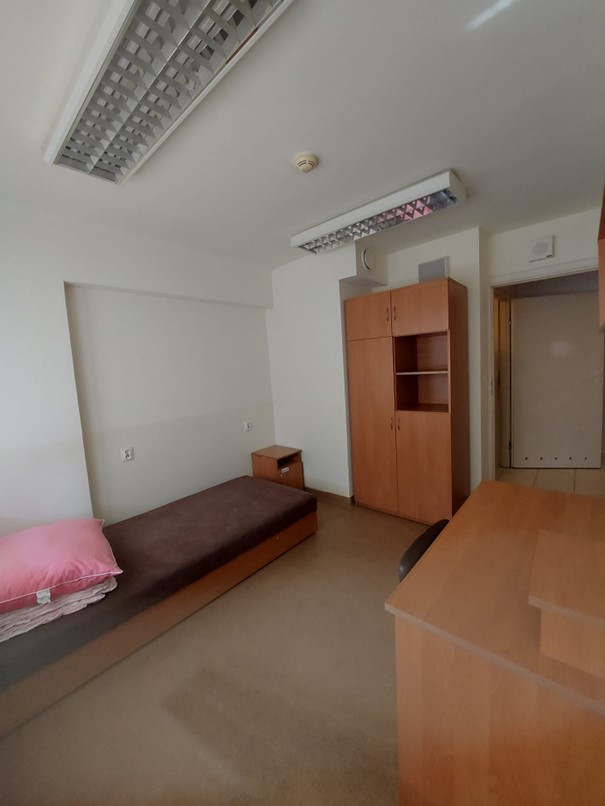 Zdjęcie przedstawia pok&oacute;j jednoosobowy w Domu Studenta Melodia. W pomieszczeniu znajduje się pojedyncze brązowe ł&oacute;żko z r&oacute;żową poduszką, biurko z krzesłem, szafa z p&oacute;łkami oraz duże okno z roletami. Pok&oacute;j jest jasny i schludny, z beżową podłogą, i drewnianymi meblami.