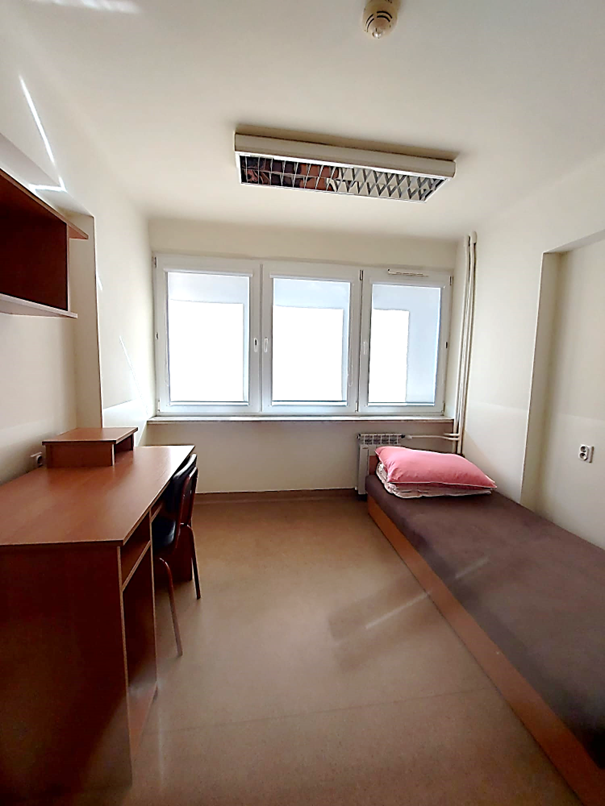 Zdjęcie przedstawia pok&oacute;j jednoosobowy w Domu Studenta Melodia. W pomieszczeniu znajduje się pojedyncze ł&oacute;żko z r&oacute;żową poduszką i białą kołdrą, biurko, krzesło oraz p&oacute;łka ścienna. Pok&oacute;j jest jasny, z beżową podłogą i ciemnobrązowymi drewnianymi meblami.