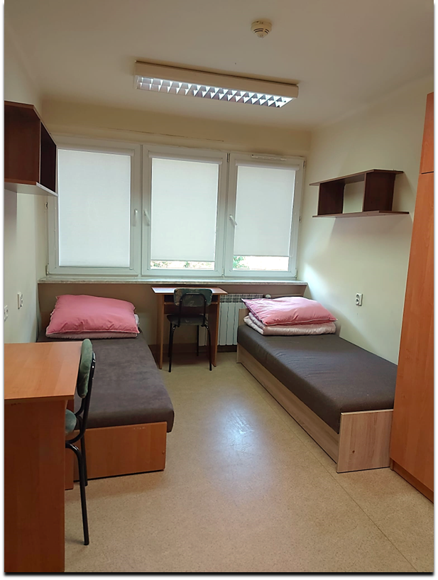 Zdjęcie przedstawia pok&oacute;j dwuosobowy w domu Studenta Melodia. W pomieszczeniu znajdują się dwa ciemnobrązowe ł&oacute;żka, dwa biurka, krzesła oraz p&oacute;łki ścienne. Pok&oacute;j jest jasny, z brązową podłogą i drewnianymi meblami.