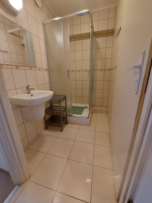 Zdjęcie przedstawia łazienkę w Domu Studenta Melodia, wnętrze wyłożone jest jasnymi płytkami. W pomieszczeniu znajduje się prysznic z przesuwnymi, matowymi drzwiami, umywalka z lustrem oraz plastikowy stojak z p&oacute;łkami. Łazienka jest czysta, dobrze oświetlona i urządzona w prostym, funkcjonalnym stylu.