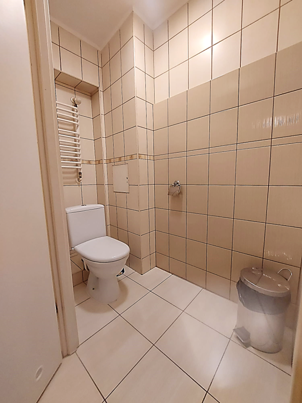 Zdjęcie przedstawia toaletę w Domu Studenta Melodia, Wnętrze wyłożone jest jasnymi beżowymi płytkami. W pomieszczeniu znajduje się kosz na śmieci, grzejnik oraz uchwyt na papier toaletowy. Łazienka jest czysta, dobrze oświetlona i urządzona w prostym, funkcjonalnym stylu.