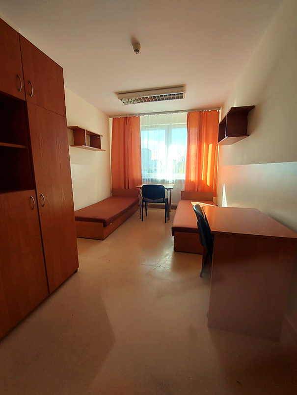 Zdjęcie przedstawia pok&oacute;j dwuosobowy w akademiku Odyseja. W pokoju znajdują się dwa pojedyncze ł&oacute;żka z brązowymi materacami, dwa biurka z krzesłami, duża szafa oraz drewniane p&oacute;łki ścienne w podobnym odcieniu. Na końcu pomieszczenia znajduje się okno zasłonięte pomarańczowymi zasłonami, przez kt&oacute;re wpada naturalne światło.