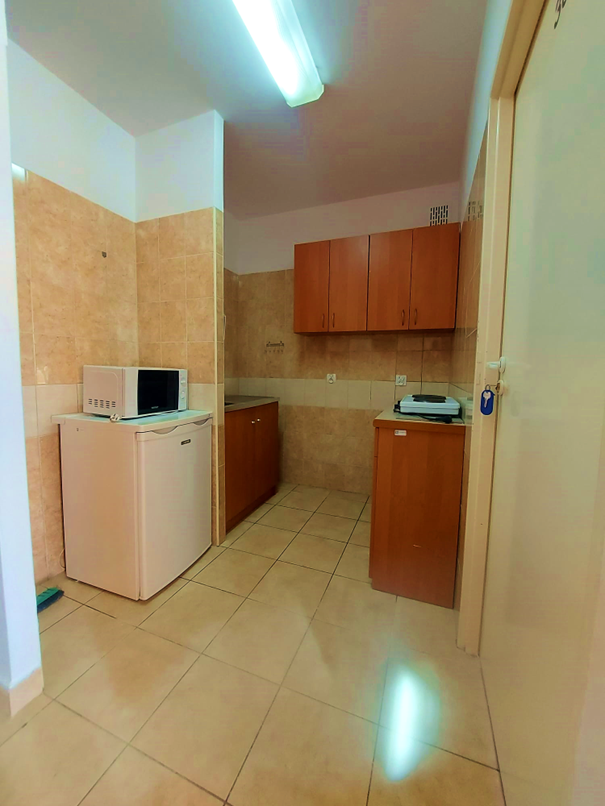 Zdjęcie przedstawia aneks kuchenny w Domu Studenta Odyseja. Pomieszczenie jest dobrze oświetlone z jasnobeżowymi płytkami na ścianach i podłodze. W pomieszczeniu znajdują się drewniane szafki, mała lod&oacute;wka z kuchenką mikrofalową oraz płyta grzewcza ustawiona na blacie. Na ścianie zamontowane są wiszące szafki w tym samym kolorze drewna.