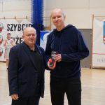 Dwaj mężczyźni na hali sportowej. Wyższy, w granatowej bluzie, trzyma nagrodę z napisem „Najlepszy Trener”. W tle zawodnicy w białych strojach koszykarskich oraz banery reklamowe.