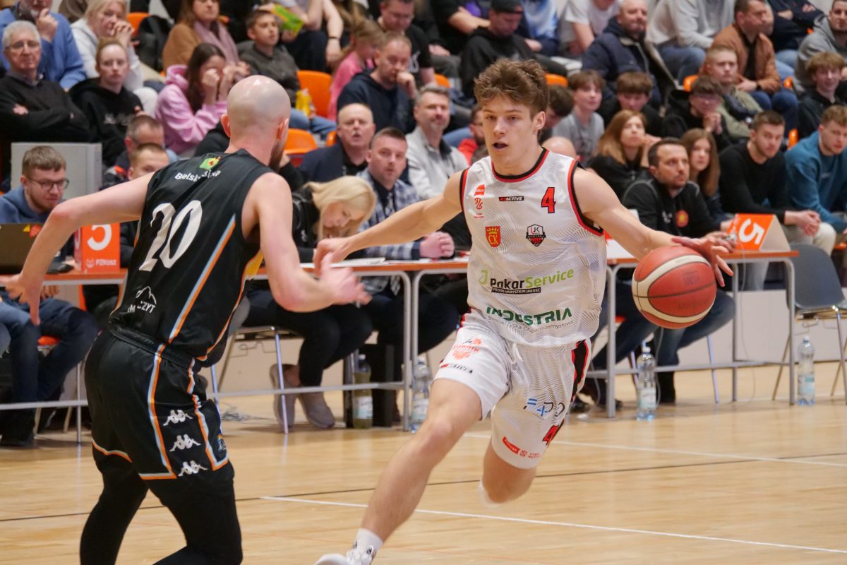 Dynamiczne ujęcie z meczu koszykówki. Młody zawodnik w białym stroju z numerem 4 drybluje piłkę, omijając przeciwnika w czarnym stroju. W tle widać trybuny wypełnione kibicami.