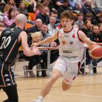 Dynamiczne ujęcie z meczu koszykówki. Młody zawodnik w białym stroju z numerem 4 drybluje piłkę, omijając przeciwnika w czarnym stroju. W tle widać trybuny wypełnione kibicami.