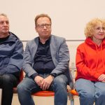 Trzy osoby siedzące na pomarańczowych krzesełkach: dwóch mężczyzn w okularach i kobieta w czerwonej bluzie z kapturem. Wyglądają na skupionych widzów lub gości wydarzenia sportowego.