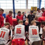 Drużyna koszykarska podczas przerwy w grze. Zawodnicy w białych koszulkach z nazwiskami (Wesołek, Cygan, Pisarczyk, Lewandowski) otaczają trenera, który udziela im wskazówek. W tle tablica wyników.