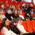 Młodzież siedzi w rzędach czerwonych foteli w auli wykładowej. Wszyscy z uwagą patrzą w jednym kierunku, słuchając prezentacji. Na pierwszym planie trzy osoby w strojach galowych i okularach.