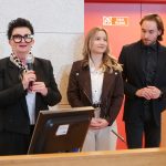 Trzy osoby na podwyższeniu. Po lewej kobieta w czarnym żakiecie i charakterystycznych okularach mówi do mikrofonu. Obok niej uśmiechnięta dziewczyna i mężczyzna w czarnym garniturze. Tło jest czerwone.