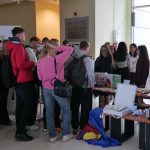 Grupa młodzieży przy stoiskach informacyjnych w jasnym holu. Na stolikach leżą gry edukacyjne, roboty i ulotki. Studenci rozmawiają z odwiedzającymi. W tle tablica informacyjna i duże okna.