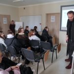 Kameralna sala wykładowa. Mężczyzna przy projektorze prezentuje urządzenie z zielonymi przyciskami. Grupa studentów siedzi w ławkach i przygląda się pokazowi. Na ekranie widoczna prezentacja.