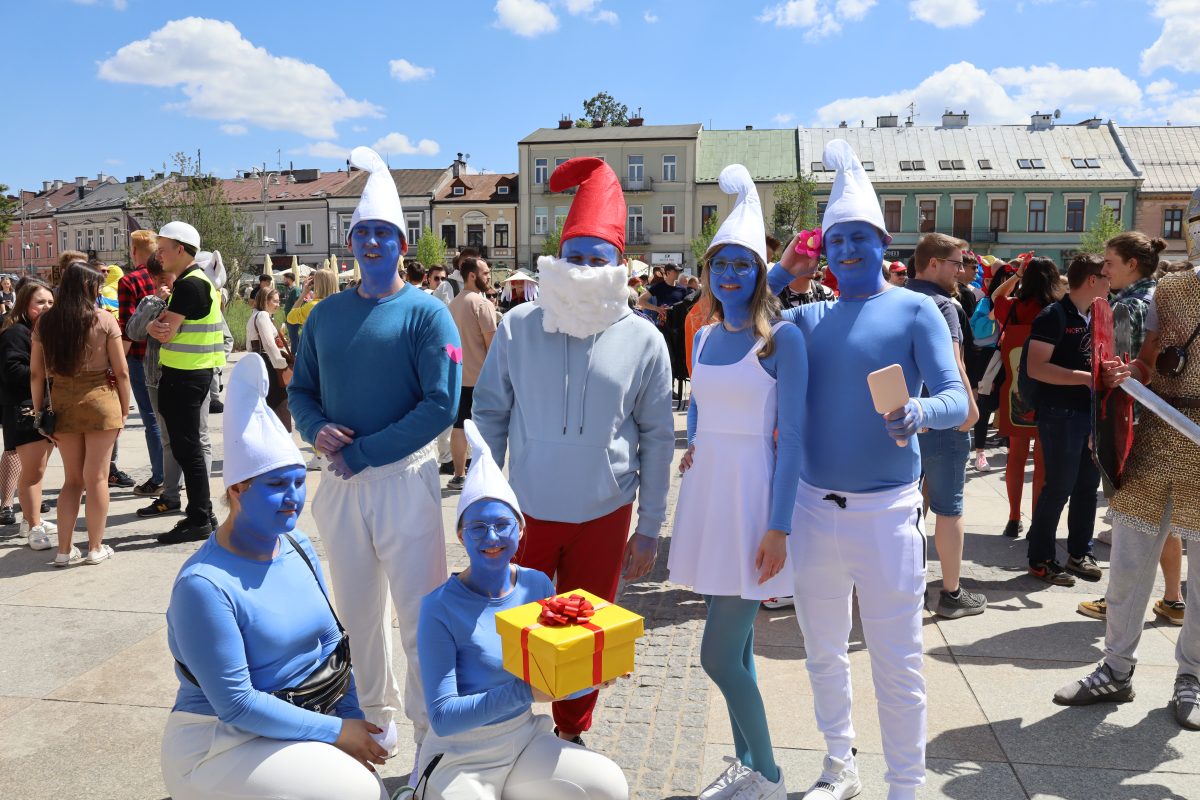 Grupa sześciu osób przebranych za Smerfy pozuje do zdjęcia na słonecznym placu miejskim. Mają niebieskie twarze i dłonie oraz charakterystyczne białe (lub czerwone w przypadku Papy Smerfa) czapki. Jedna osoba trzyma żółty prezent z czerwoną kokardą. W tle widać kolorowe kamienice i tłum ludzi w różnych strojach. Atmosfera jest radosna i piknikowa.