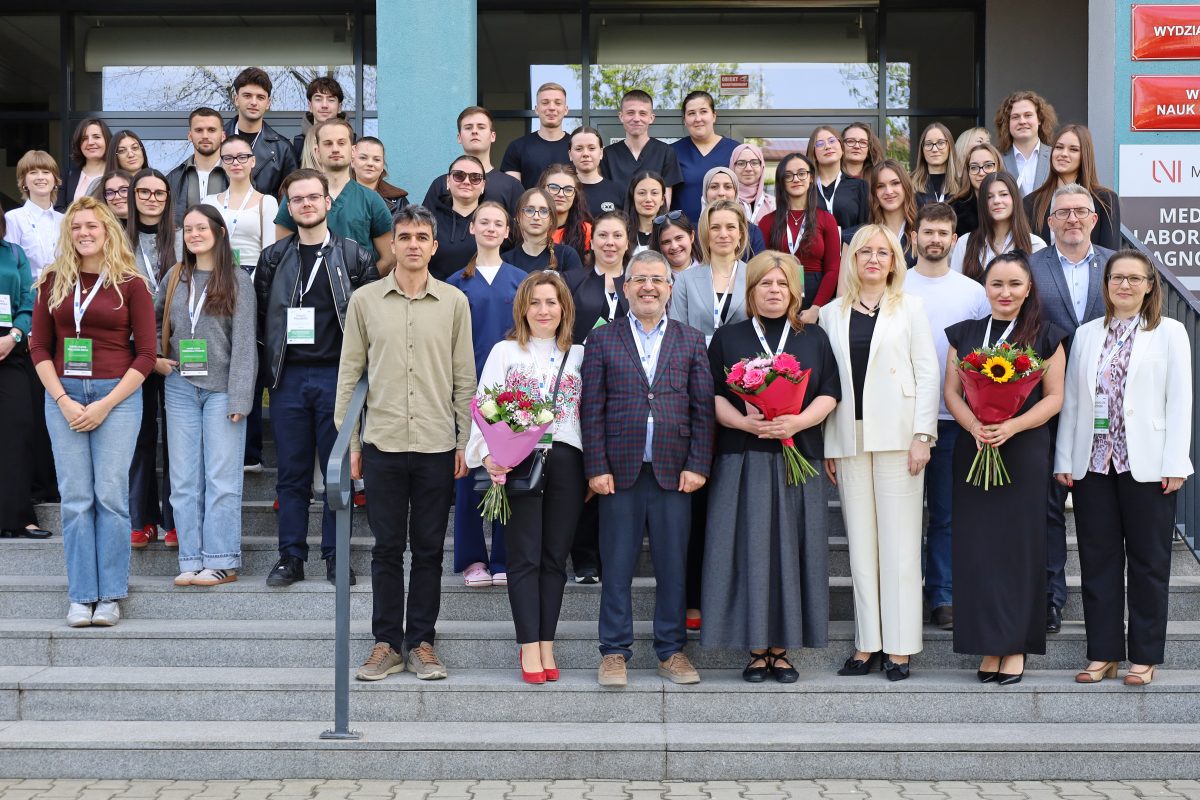 Grupa kilkudziesięciu studentów i wykładowców pozuje na schodach przed budynkiem Collegium Medicum UJK. W pierwszym rzędzie osoby trzymające bukiety kwiatów. Nad wejściem widoczne tablice „Wydział Lekarski” i „Wydział Nauk o Zdrowiu”.