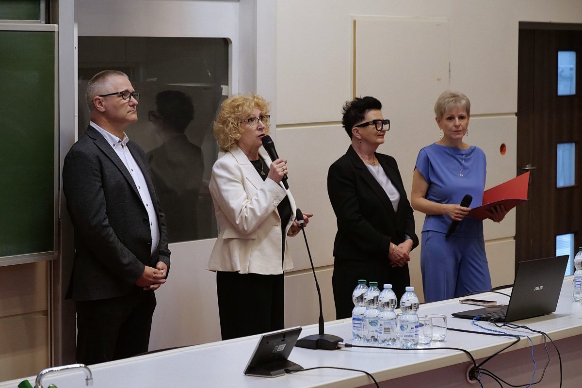 Cztery osoby stojące za stołem prezydialnym w auli. Kobieta w białej marynarce mówi do mikrofonu. Obok niej stoją: mężczyzna w ciemnej marynarce oraz dwie kobiety – jedna w czarnym żakiecie, druga w niebieskiej bluzce z czerwoną teczką. Na stole widać laptopa, butelki z wodą i szklanki.