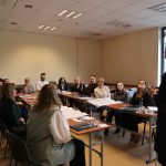 Uczestnicy szkolenia siedzą przy stołach ustawionych w podkowę i słuchają prelegentki, kt&oacute;ra stoi tyłem do obiektywu po prawej stronie. Na stołach widać notatniki, arkusze papieru i filiżanki kawy.