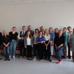 Oficjalne zdjęcie grupowe uczestnik&oacute;w szkolenia (kobiety i mężczyźni) stojących pod białą ścianą w sali wykładowej. Uczestnicy są uśmiechnięci, ubrani w stroje biznesowe i casualowe.