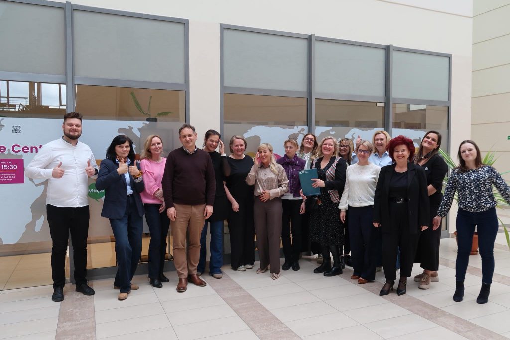 Zdjęcie grupowe w holu: Grupa uczestnik&oacute;w projektu pozuje wsp&oacute;lnie w jasnym, przeszklonym holu. Część os&oacute;b uśmiecha się i pokazuje kciuki w g&oacute;rę, w tle widoczny jest zarys mapy świata na szybie.