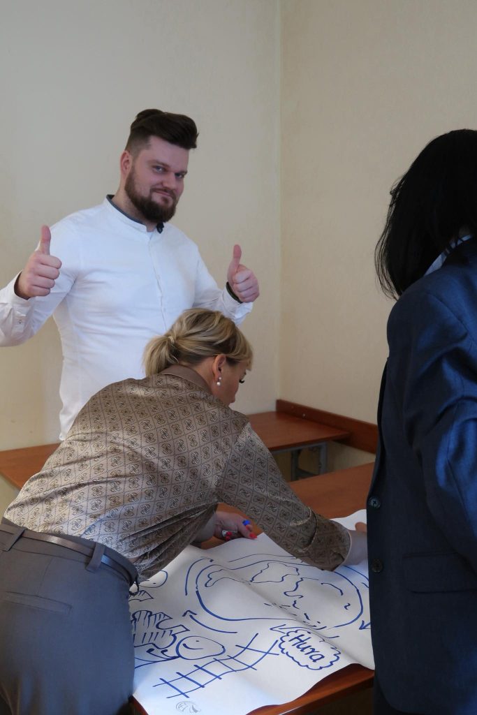 Rysowanie na arkuszu: Mężczyzna pokazuje kciuki w g&oacute;rę, podczas gdy obok niego uczestniczka nanosi kolorowe rysunki na duży arkusz papieru.