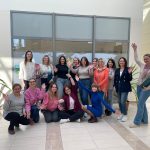 Grupa kilkunastu os&oacute;b pozuje do radosnego zdjęcia w przeszklonym atrium. Część os&oacute;b kuca, inne stoją i machają do aparatu. W tle na szybie widoczna naklejka z mapą świata i napisem "Welcome"