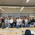 Zdjęcie grupowe kilkunastu kobiet stojących w bibliotece na tle regał&oacute;w wypełnionych książkami. Przed nimi znajduje się długi st&oacute;ł konferencyjny z rozłożonymi materiałami szkoleniowymi