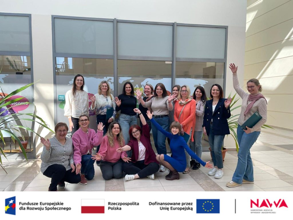 Zdjęcie grupowe (hol): Radosne zdjęcie grupowe uczestniczek projektu w przeszklonym holu. Kobiety uśmiechają się i machają do aparatu, a w tle na szybie widać zarys mapy świata.