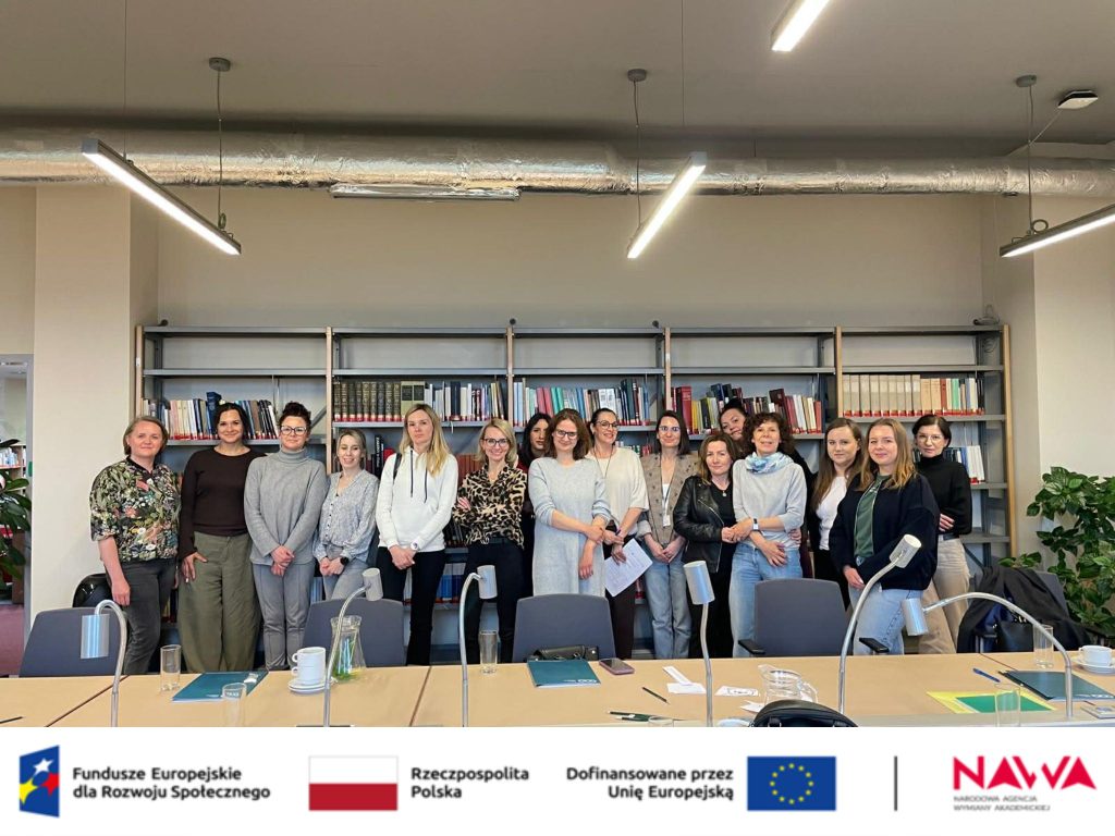 Zdjęcie grupowe (sala): Pozowane zdjęcie grupowe uczestniczek szkolenia. Kobiety stoją uśmiechnięte w rzędzie na tle wysokich regał&oacute;w bibliotecznych w jasnym pomieszczeniu.