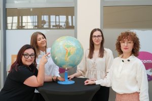 Cztery uśmiechnięte kobiety w eleganckich strojach stoją wokół wysokiego stolika z globusem na tle przeszklonego wejścia do budynku.