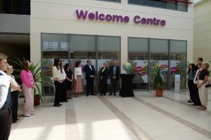 Grupa kilkunastu osób uczestniczy w oficjalnym wydarzeniu w jasnym, przestronnym holu przed wejściem do „Welcome Centre”.