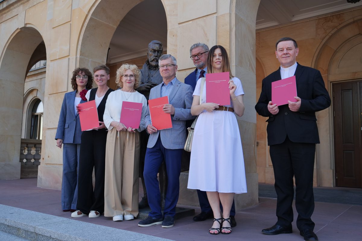 Grupa sześciu elegancko ubranych osób stoi przed zabytkowym budynkiem z arkadami, obok pomnika. Cztery kobiety i dwóch mężczyzn trzymają w dłoniach czerwone teczki z białym godłem i napisami. Wszyscy uśmiechają się w stronę obiektywu.