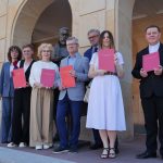 Grupa sześciu elegancko ubranych osób stoi przed zabytkowym budynkiem z arkadami, obok pomnika. Cztery kobiety i dwóch mężczyzn trzymają w dłoniach czerwone teczki z białym godłem i napisami. Wszyscy uśmiechają się w stronę obiektywu.