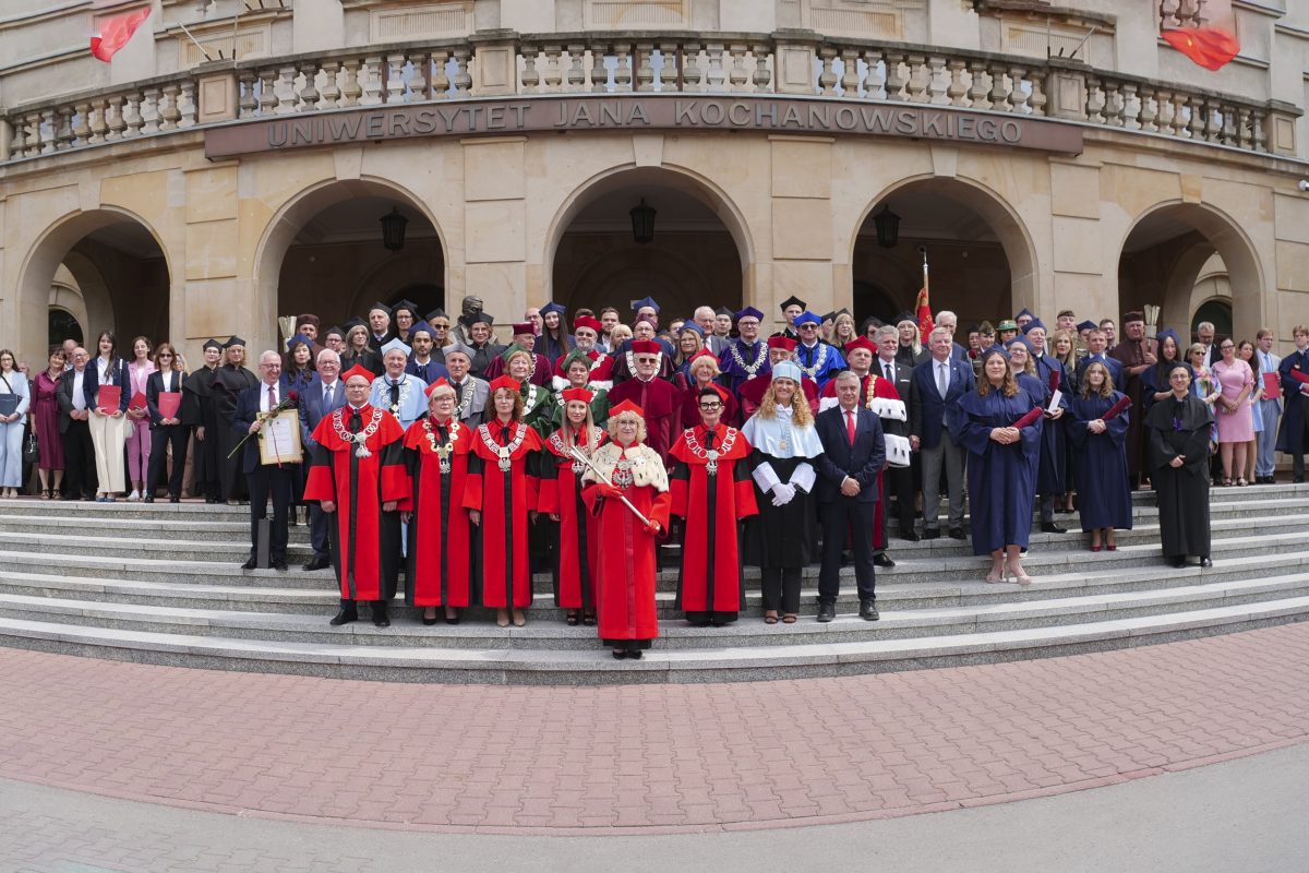 Grupowe zdjęcie uczestników uroczystości akademickiej wykonane przed głównym wejściem do budynku Uniwersytetu Jana Kochanowskiego w Kielcach. Na schodach stoją osoby w togach i strojach ceremonialnych oraz zaproszeni goście.